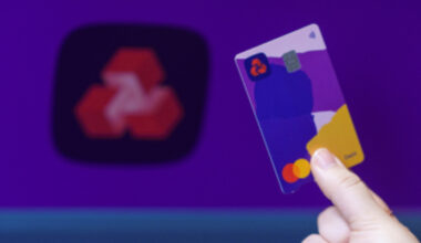 Hand holding a NatWest debit card with a colorful design, blurred NatWest logo in the background.