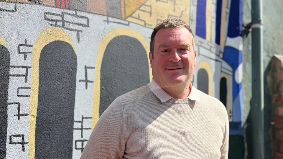 Head and shoulders image of Ryan Coogan standing in front of a colourful wall. He has short brown hair and is wearing a beige jumper with a light shirt underneath.