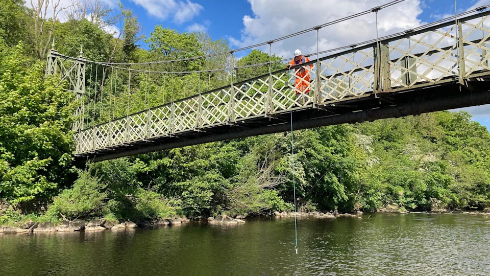 A man lowers a small bucket off a bridge above the River Wharfe at Ilkley so that he can test water pollution levels. The bucket is suspended on a rope.