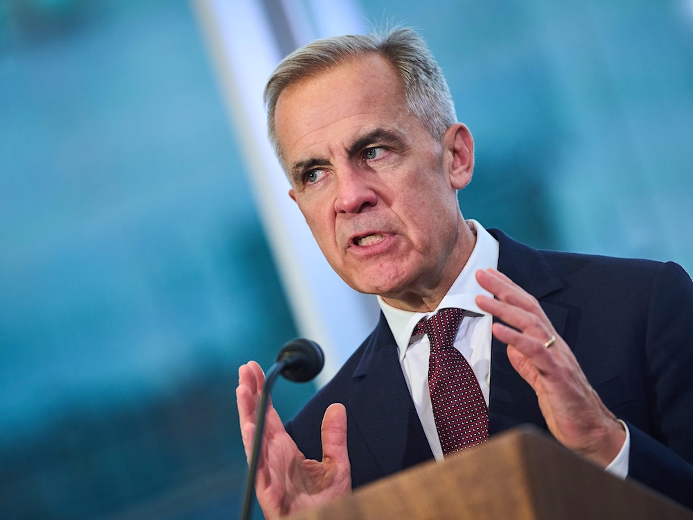 A man in his 60s with light skin and short grey hair speaks at a lectern. The photograph is tilted on an angle.