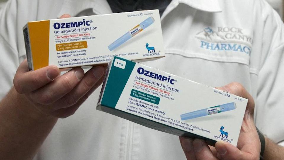 A pharmacist holding two boxes in each hand of Ozempic, showing different doses (one 2mg and another 1mg). The boxes say Ozempic (semaglutide injection) and feature a photo of the inkection pen, as well as instructions on how to use the drug. They also feature the logo of manufacturer Novo Nordisk.