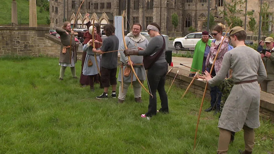 A line of people are standing in a grassed area while holding medieval-style bows and arrows, with people dressed in medieval costume guiding them. Several people nearby are taking photos and a grand historical building can be seen in the background.
