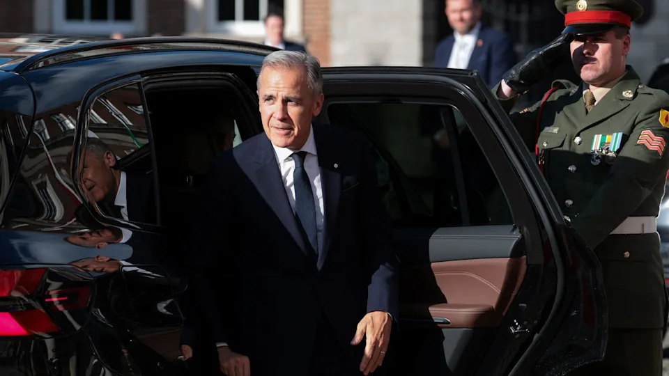 Carney steps out of a shiny black car. He wears a sharp suit. Behind him a solider salutes.