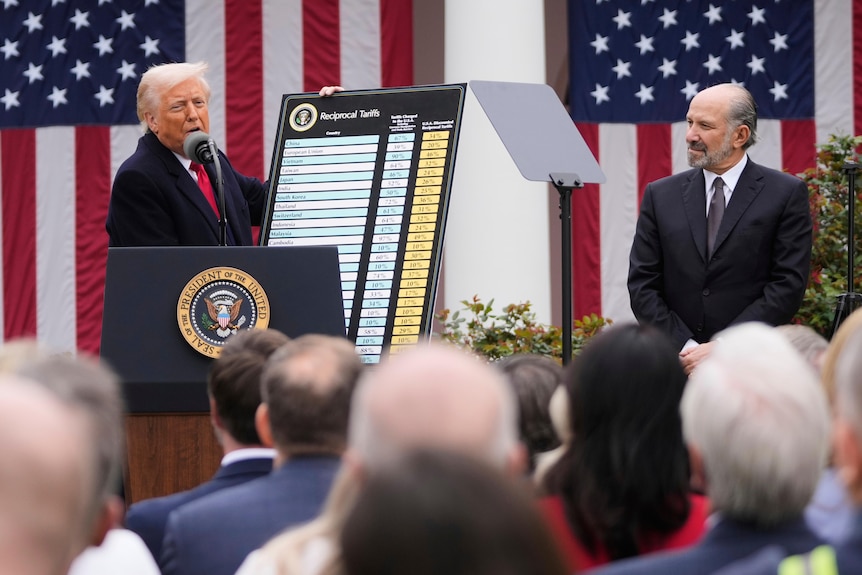 Donald Trump stands on stage in front of American flags, holding a large board with a list on it.