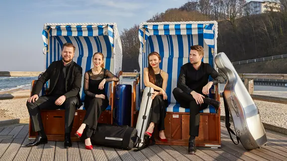 Vier Musiker*innen sitzen in Konzertkleidung verteilt in zwei Strandkörben, vor sich ihre Instrumentenkoffer | Oliver Borchert Vier Musiker*innen sitzen in Konzertkleidung verteilt in zwei Strandkörben, vor sich ihre Instrumentenkoffer
