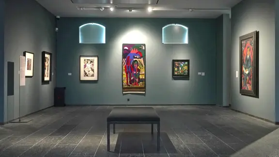 Durch die Position zueinander, werden die Kunstwerke in einen Dialog miteinander gesetzt. | Screenshot Blick in den Ausstellungsraum mit mehreren beleuchteten Gemälden