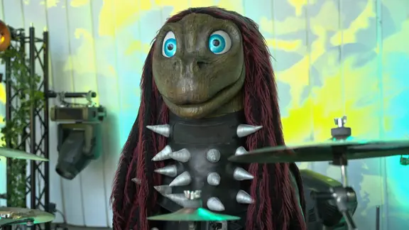 Ein als Dino verkleideter Mensch mit großen blauen Augen sitzt vor einem Schlagzeug | NDR Ein als Dino verkleideter Mensch mit großen blauen Augen sitzt vor einem Schlagzeug