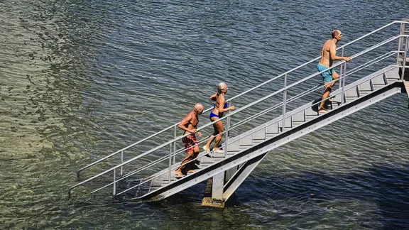 Menschen in Badeanzügen steigen eine Metalltreppe aus dem Wasser hinauf an einem sonnigen Tag. | IMAGO / imagebroker Menschen in Badeanzügen steigen eine Metalltreppe aus dem Wasser hinauf an einem sonnigen Tag.
