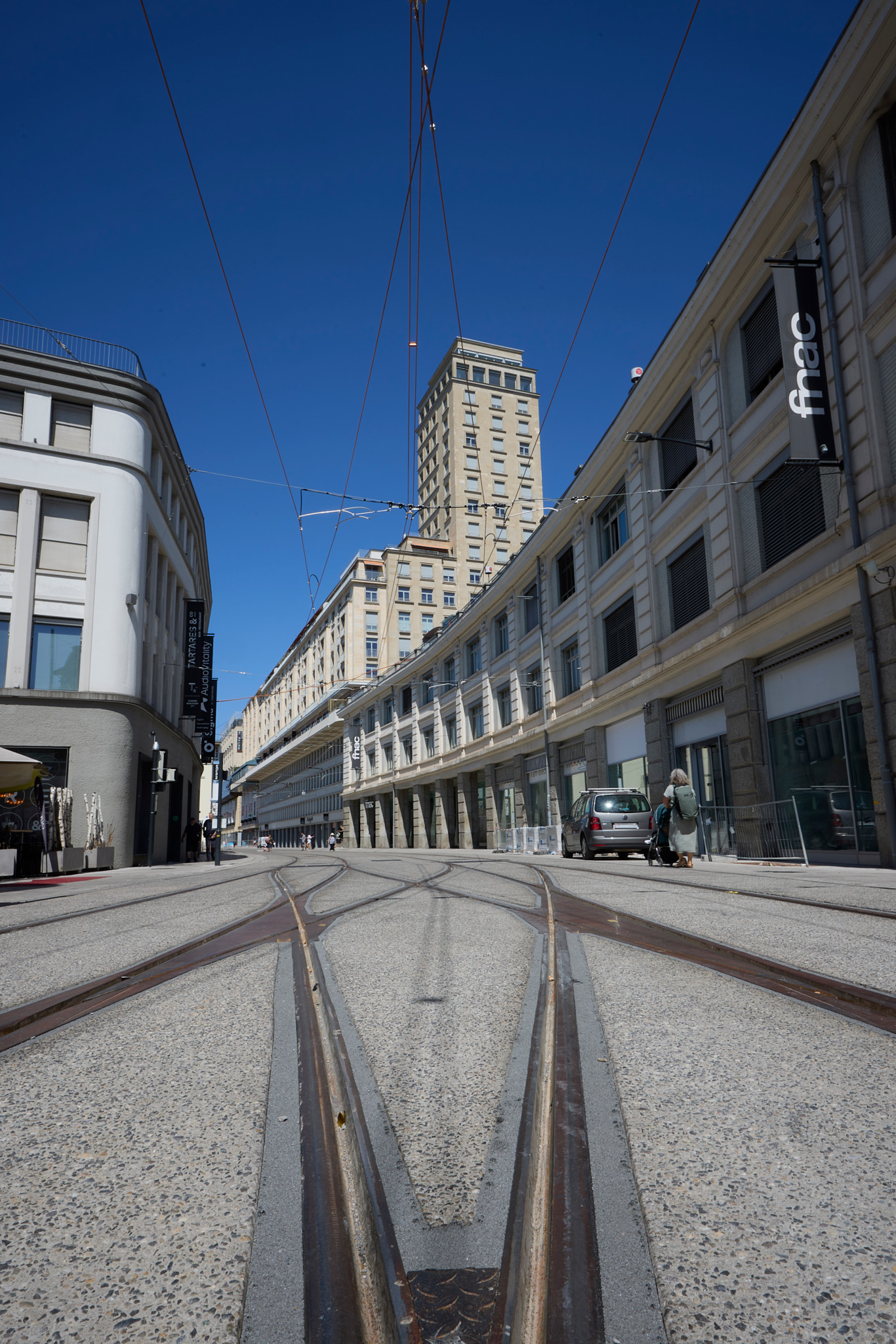 Rails du futur tram Lausanne-Flon–Renens rue de Genève, sans arbres, entre le MAD et la Fnac, sous un ciel bleu.