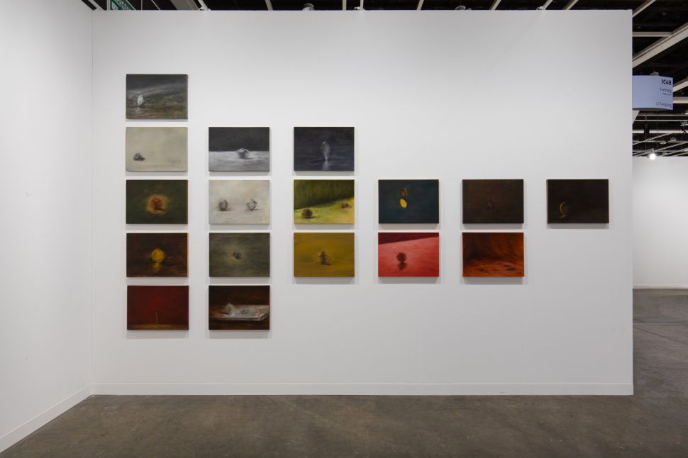 A wall installation featuring a series of small, atmospheric oil paintings arranged in a grid pattern. The paintings, varying in tones from dark and moody to vibrant reds and yellows, depict abstract forms with subtle, softly glowing details, creating an ethereal and contemplative visual experience.