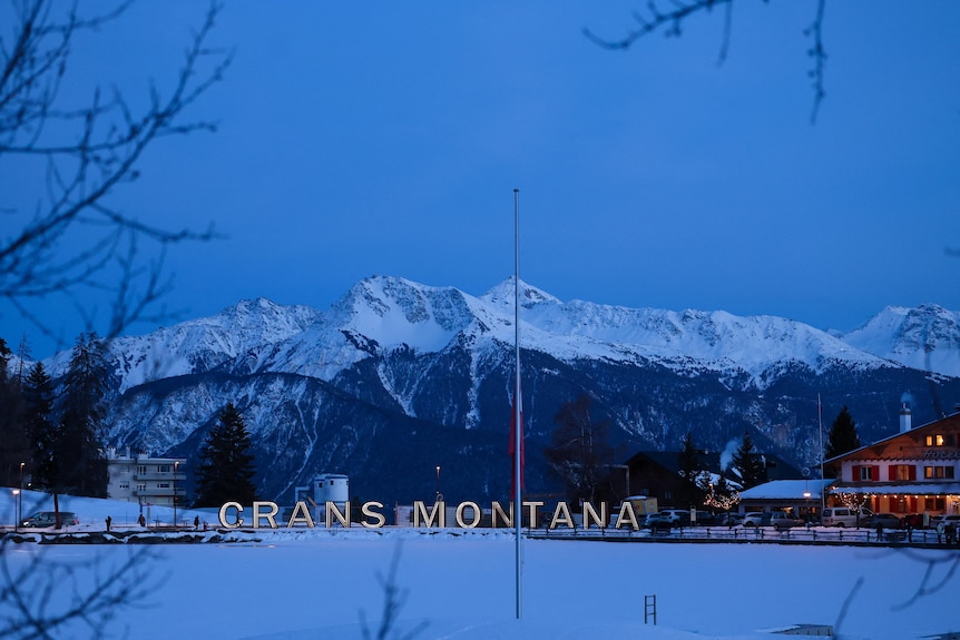 Illuminated letters spelling out the name Crans‑Montana stand behind a flag at half‑mast