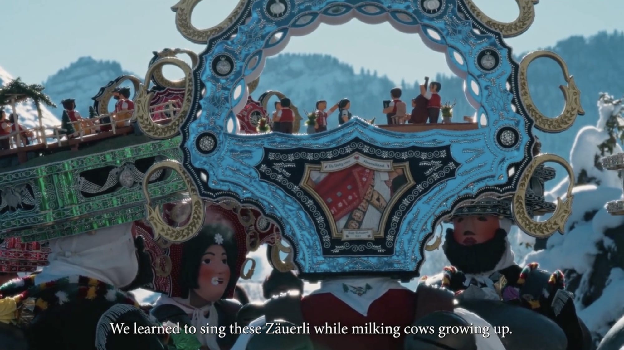 A still from a short film about the Silvesterchlausen tradition in Switzerland featuring men wearing elaborate costumes and headdresses. Text at the bottom reads,