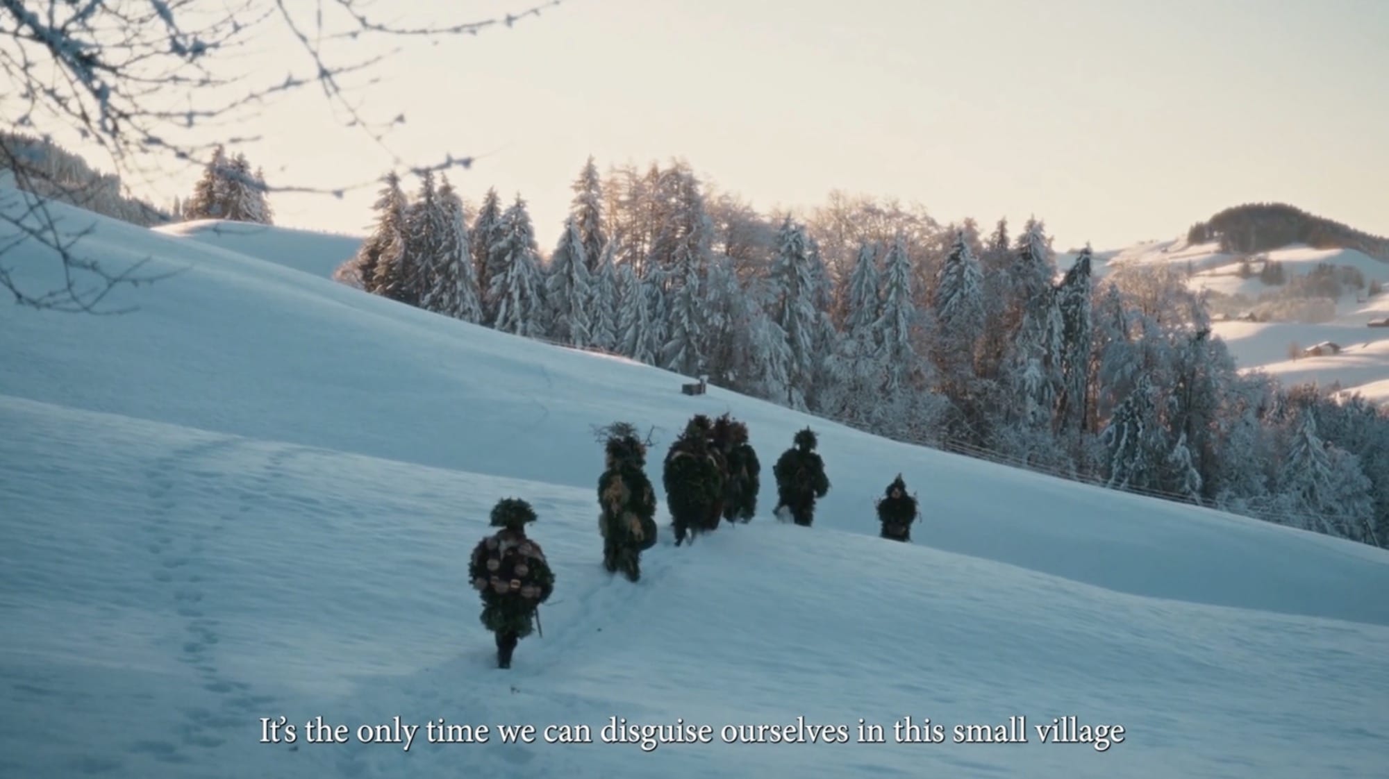 A still from a short film about the Silvesterchlausen tradition in Switzerland showing a line of costumed men walking across a snowy hill. Text on the bottom reads, "It's the only time we can disguise ourselves in this small village"