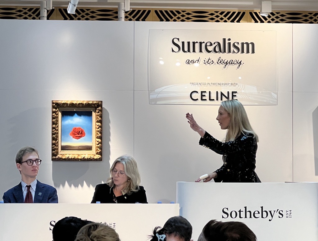 Sotheby’s auctioneer conducting the ‘Surrealism and Its Legacy’ sale in partnership with Celine, with a surrealist painting of a floating red rose displayed on the wall behind.