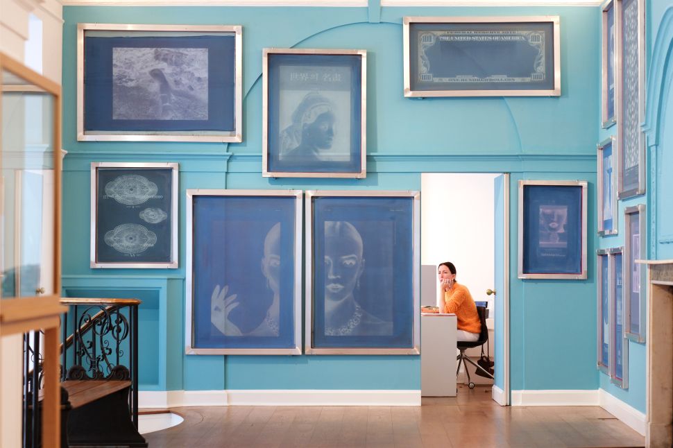 An aqua-blue gallery room displays framed blue-toned works across the walls, with a person seated at a desk visible through an open doorway.