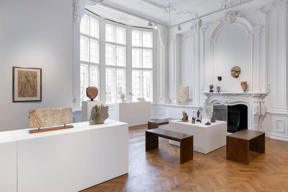 An ornate white gallery room with tall windows, parquet floors and decorative molding displays small sculptures, masks and reliefs on white plinths and benches around a carved fireplace.
