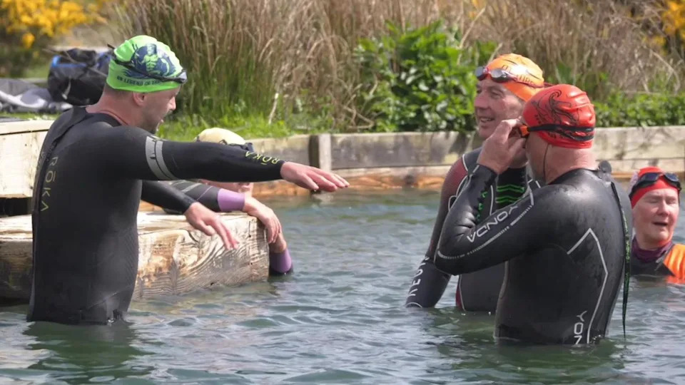 Neil standing in the water in a wet suit, with his arm outstretched showing his swimming technique to two other swimmers in wet suits
