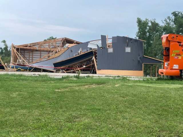 A metal building has collapsed, with its roof and walls partially fallen. Construction equipment is visible nearby on a grassy area.