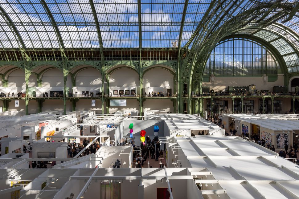 A crowded view of Art Basel Paris inside the Grand Palais, with visitors moving between gallery booths beneath the building’s vast glass-and-iron roof.