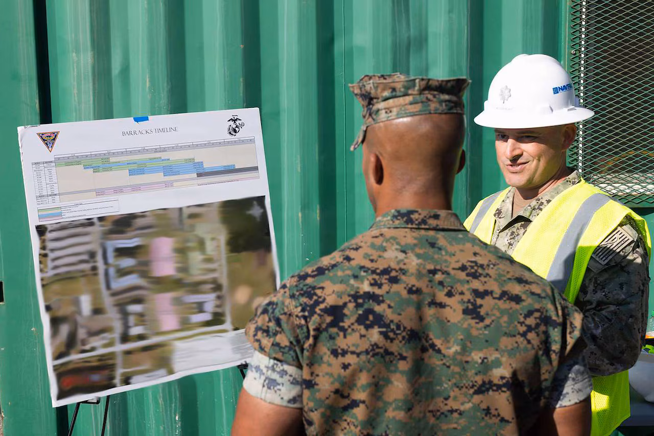 A man in a camouflage military uniform, safety vest and hard hat talks to another man in a camouflage military uniform outside as they look at an architectural rendering on an easel.