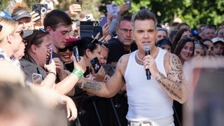 Robbie Williams während eines Konzert im Freien an einer Absperrung, wo er von vielen Fans fotografiert wird.