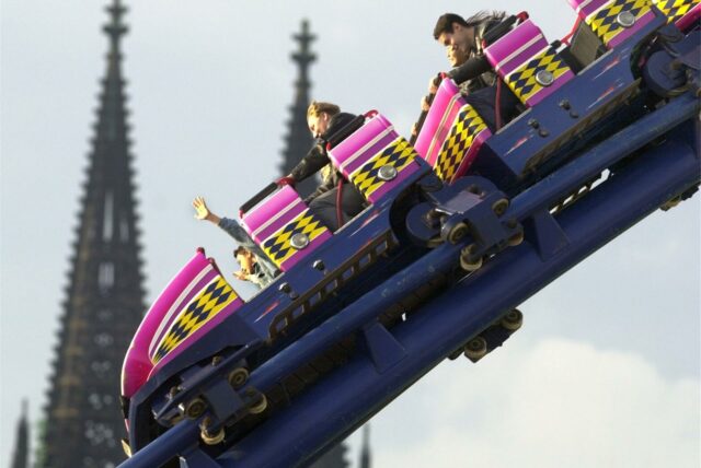 Besucher sitzen auf der Deutzer Kirmes in Köln in einer Achterbahn, während im Hintergrund die Türme des Kölner Doms zu sehen sind.