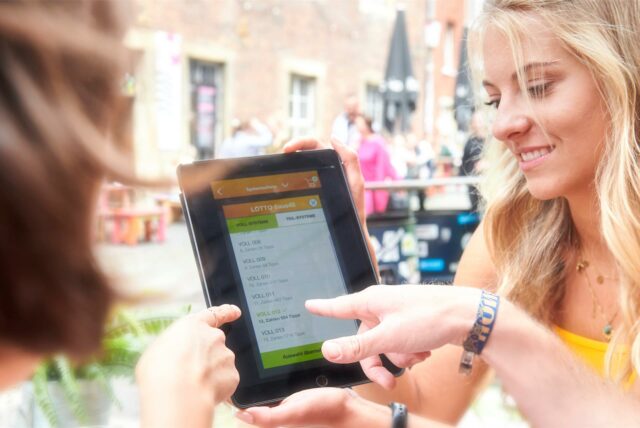 Zwei Personen betrachten ein Tablet mit einer Lotto 6aus49-App. Auf dem Bildschirm sind verschiedene Systemscheine zur Auswahl dargestellt.
