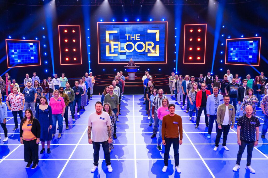 Die 100 Quizzer stehen aufgereiht in ihren Quadraten für ein Foto bereit. im Hintergrund ist in goldener Schrift auf blauem Untergrund 