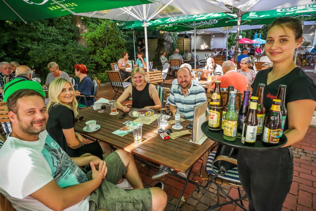 Eine Kellnerin steht lächelnd mit einem Tablet voller Getränke an einem Biergarten Tisch, an dem lachende Gäste sitzen.