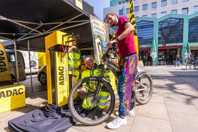Ein Mann am ADAC-Stand des E-Bike-Festivals in Dortmund lächelt in die Kamera und hält sein Rad fest. Am Vorderreifen kniet ein Mitarbeiter vom ADAC-Stand
