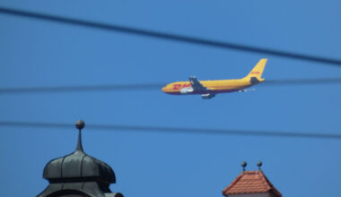 DHL-Flugzeug am Himmel. Dazu Oberleitungen und Türmchen.