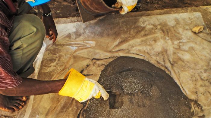 Hand mit gelbem Arbeitshandschuh schaufelt einen kleinen Berg dunkelbraunen Erzes um