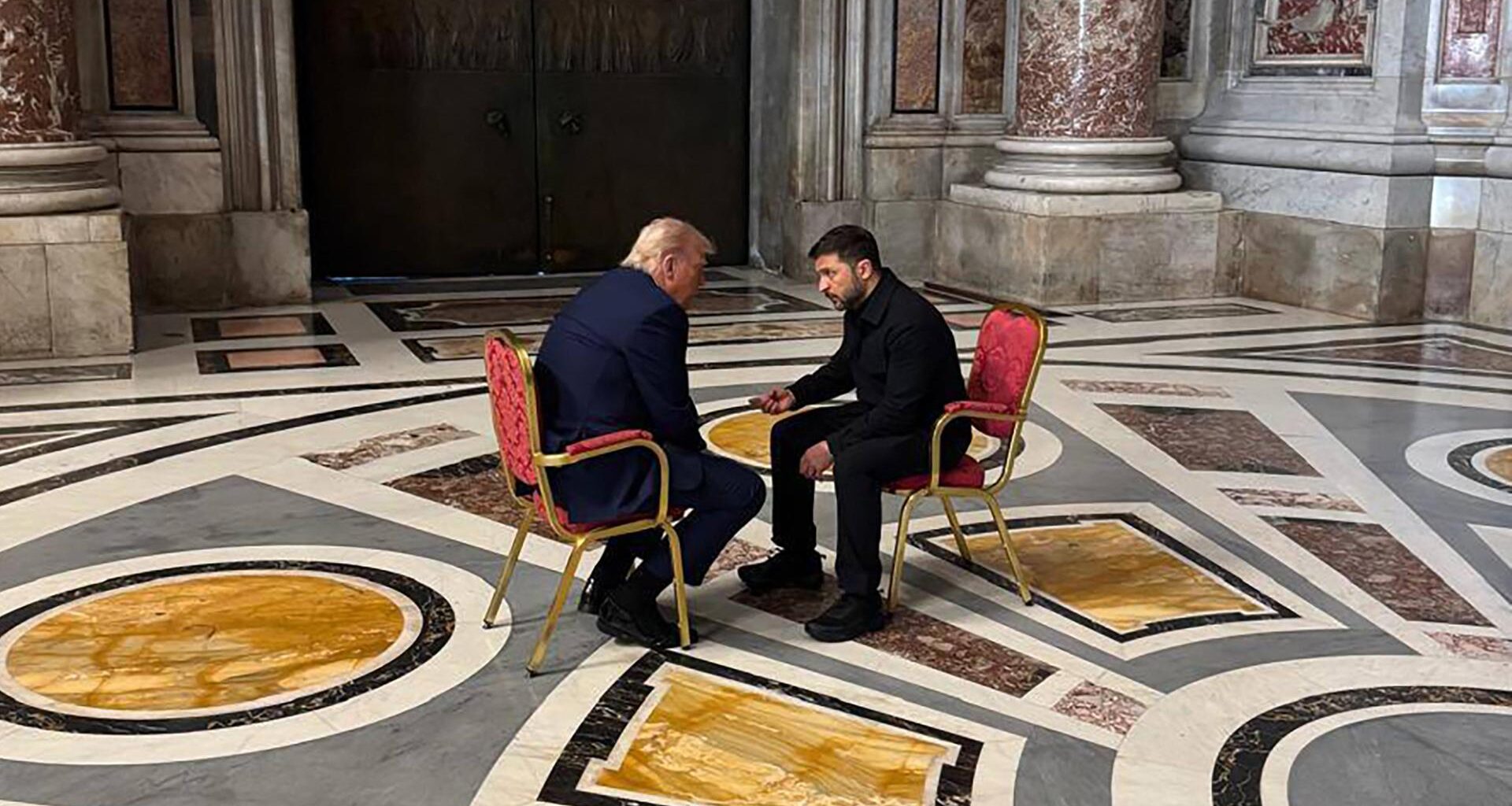 Das Foto zeigt US-Präsident Trump und den ukrainischen Präsidenten Selenskyj im Gespräch miteinander. Beide sitzen sich gegenüber auf goldenen Stühlen, die mit rotem Stoff bezogen sind.