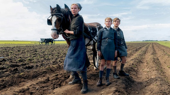 Eine Frau mit Kopftuch steht neben einem Pferd, das vor einen Pflug angespannt ist und schaut nervös, neben ihr auf dem Acker stehen zwei Jungen  - Filmszene mit Diane Kruger aus dem Film "Amrun" © 2025 bombero international GmbH & Co. KG / Rialto Film GmbH / Warner Bros. Entertainment GmbH / Mathias Bothor Foto: Mathias Bothor