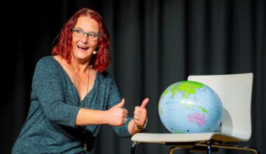 Eine Frau mit roten Haaren und Headset-Mikrofon gestikuliert lächelnd auf einer Bühne. Sie kniet vor einem Stuhl, auf dem ein aufgeblasener Globus liegt und streckt die Daumen nach oben.