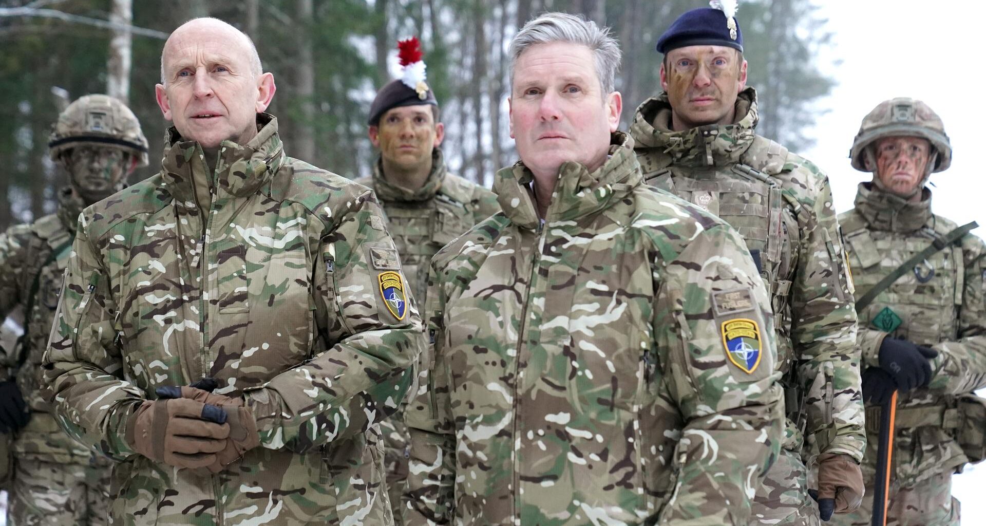Keir Starmer und John Healey stehen mit vier Soldaten des britischen Militärs in militärischen Uniformen vor einem Wald.