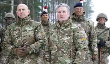 Keir Starmer und John Healey stehen mit vier Soldaten des britischen Militärs in militärischen Uniformen vor einem Wald.