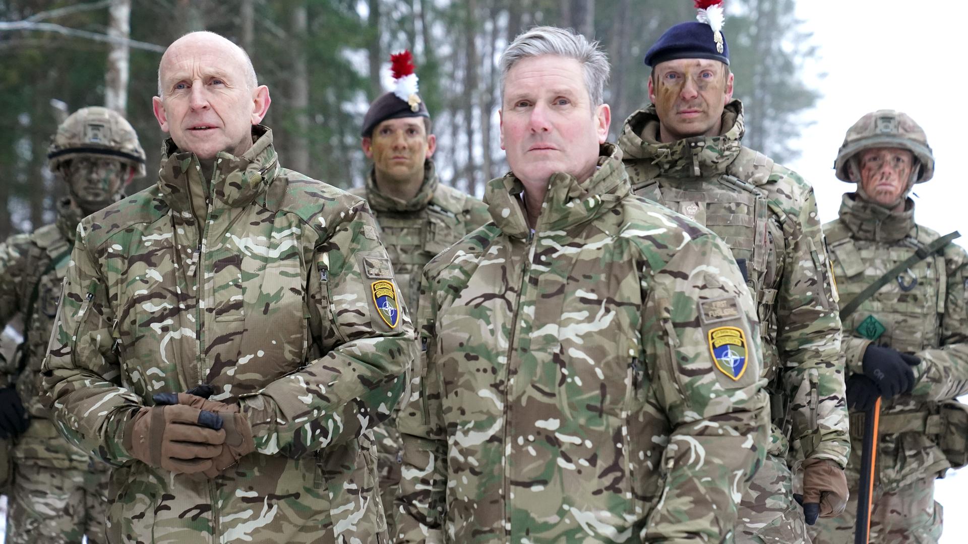 Keir Starmer und John Healey stehen mit vier Soldaten des britischen Militärs in militärischen Uniformen vor einem Wald.