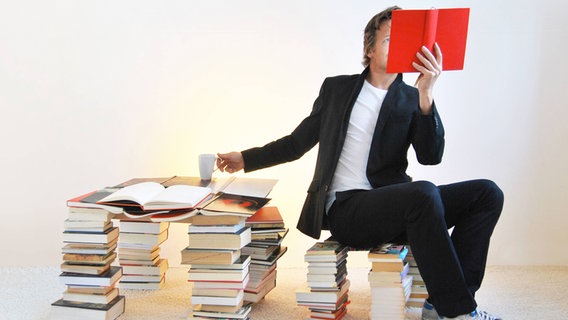 Ein Mann sitzt auf einem Bücherstapel und liest ein rotes Buch. © Imago Images Shotshop 