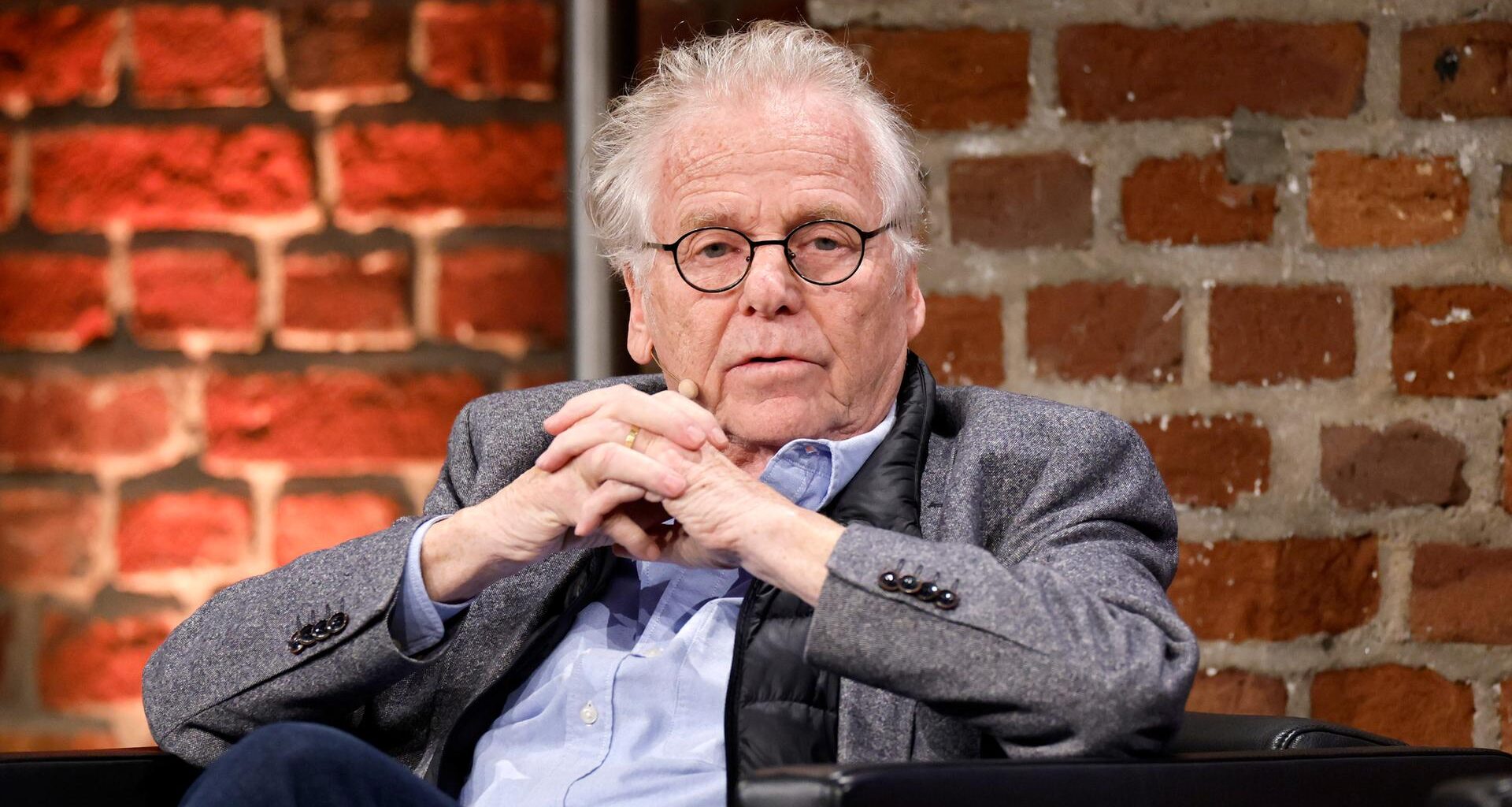 Der frühere Grünen-Politiker und Publizist Daniel Cohn-Bendit (Grüne) im grauen Jacket, hält die Hände verschränkt und sitzt vor einer Ziegelsteinwand.