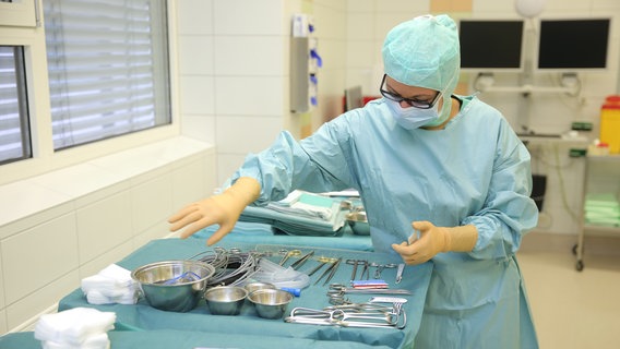 In einem Operationssaal steht eine Person im Schutzanzug mit Mundschutz vor einem Tisch mit Operationsbesteck. © Diako Flensburg 