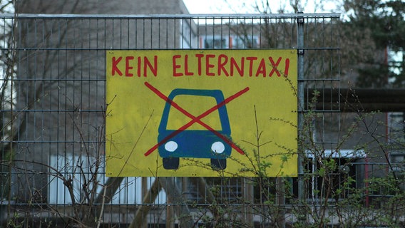 Am Zaun einer Schule hängt ein Schild mit einem durchgestrichenen Auto und der Aufschrift "Elterntaxi". © IMAGO Foto: Hanno Bode