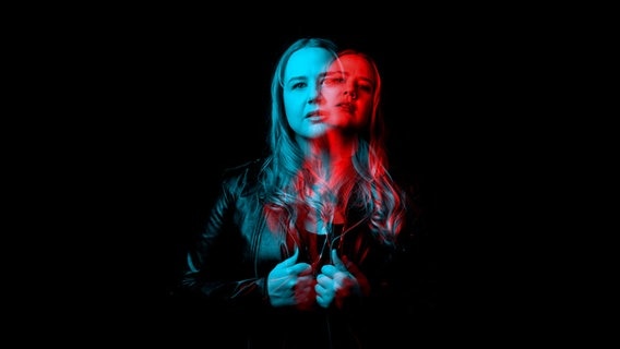 Plakatmotiv der Staatsoper Hannover für "The Greek Passion". Das Bild zeigt eine Frau mit langen Haaren, die eine schwarze Lederjacke trägt und den Jackenrevers in beiden Händen hält. Der Hintergrund ist dunkel, und es gibt einen blauen und roten Lichteffekt auf der Person, der eine Art 3D-Effekt erzeugt. © Staatsoper Hannover Foto: Clemens Heidrich