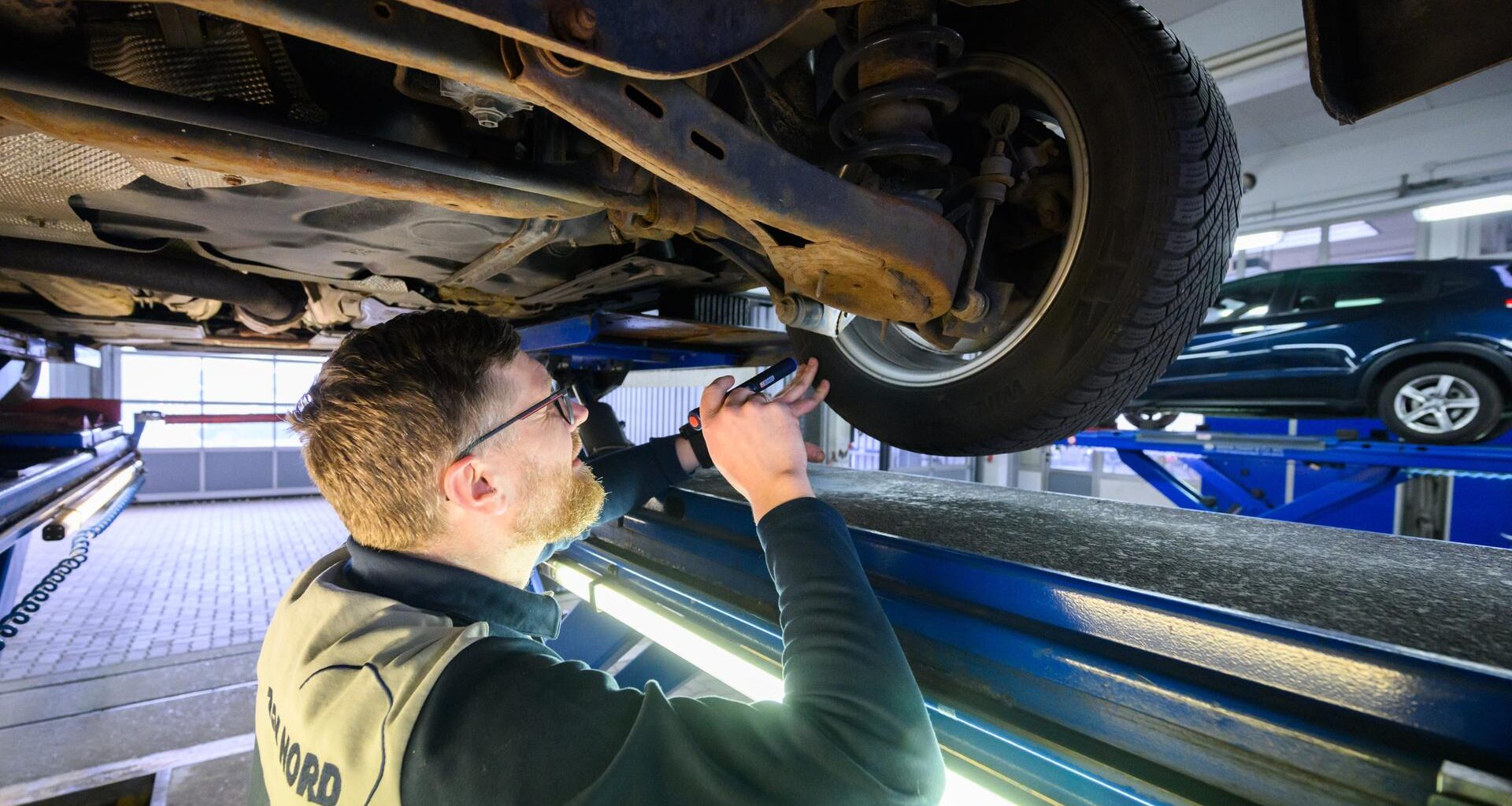 Ein Sachverständiger untersucht in einer Werkstatt die Unterseite eines hochgebockten Fahrzeugs mit einer Taschenlampe.