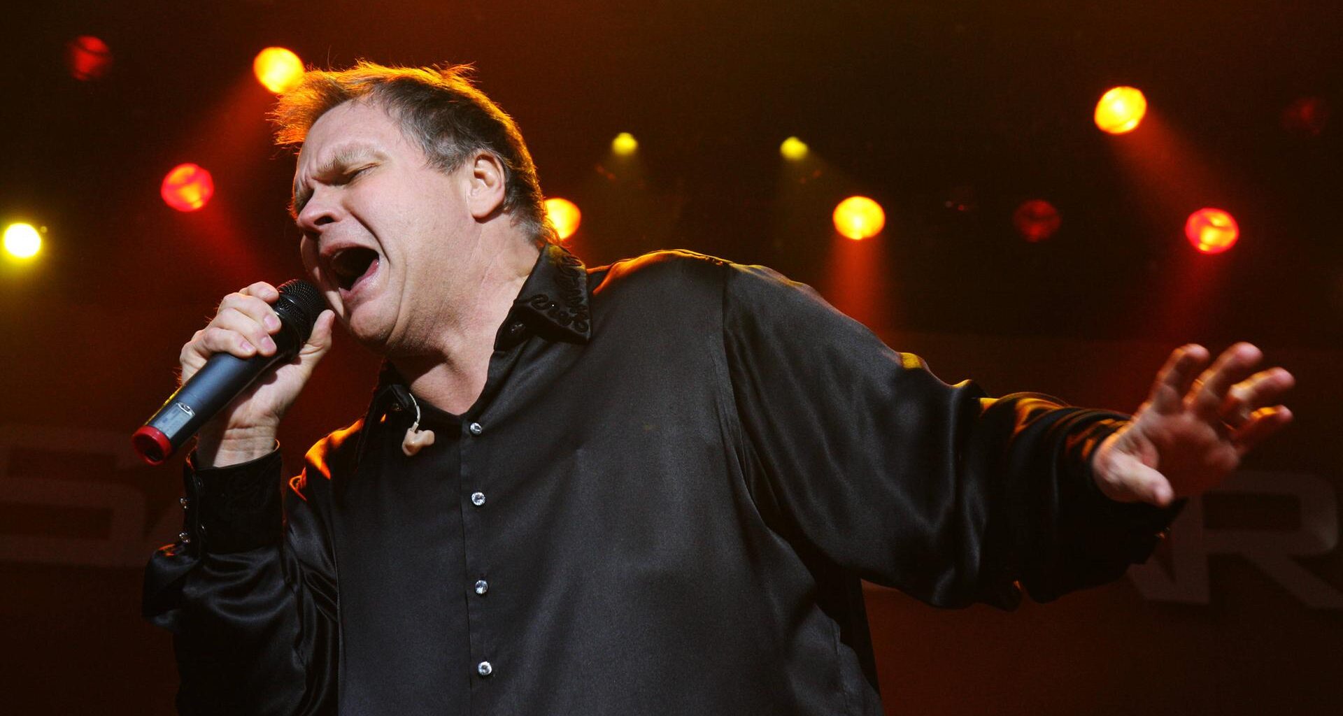 Ein Mann steht mit ausgebreiteten Armen auf einer Bühne und singt. In der rechten Hand hält er ein Mikrofon. Es ist der US-Rockmusiker Meat Loaf.