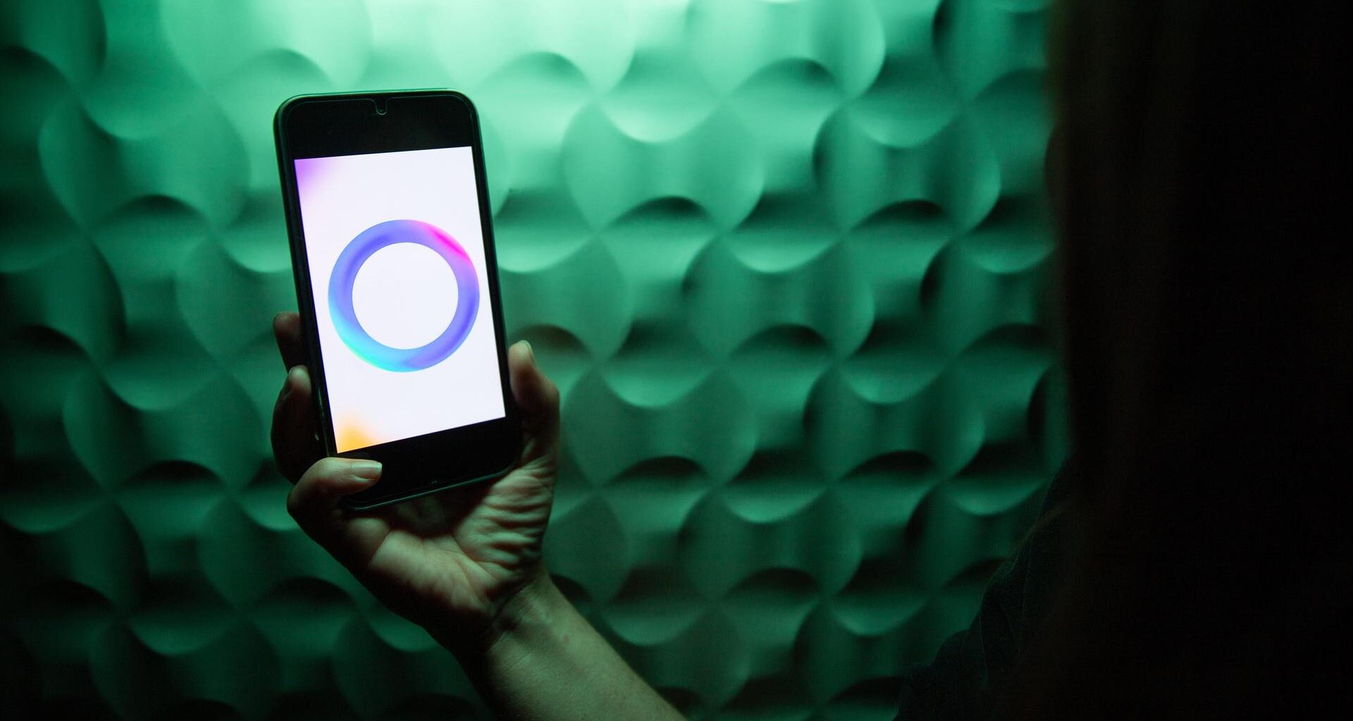 Eine Hand hält ein Smartphone, auf dem das Meta-Logo für ihre KI angezeigt wird: Ein Ring aus ineinanderfließenden Farben in Blau, Lila und Pink.