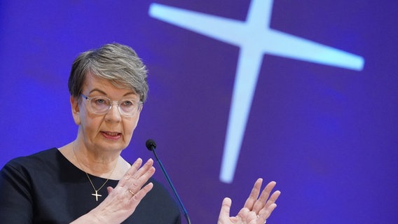 Landesbischöfin Kristina Kühnbaum-Schmidt steht auf einer Konferenz vor einem blauen Hintergrund, auf dem ein weißes Kreuz zu sehen ist. © picture alliance Foto: Marcus Brandt
