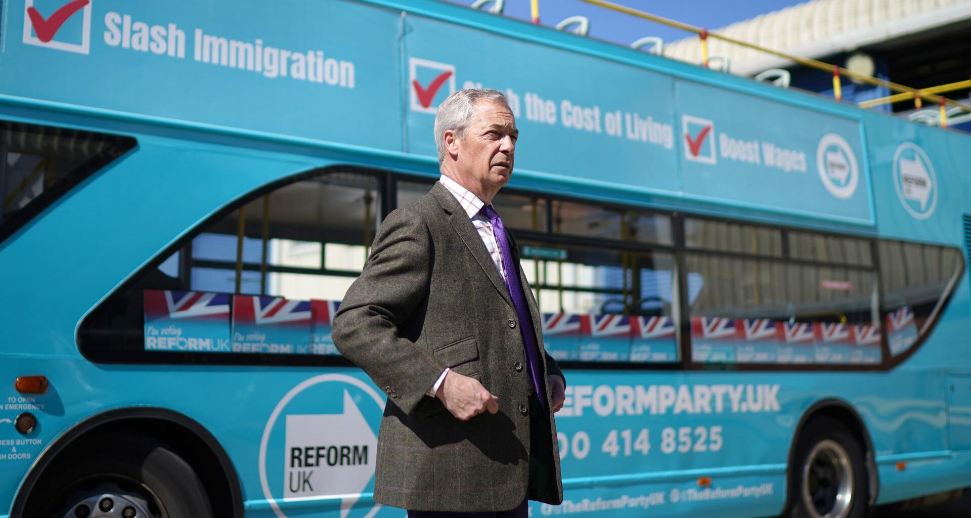 Nigel Farage steht während seiner Wahlkampftour vor einem hellblauen Werbebus seiner Partei Reform UK.