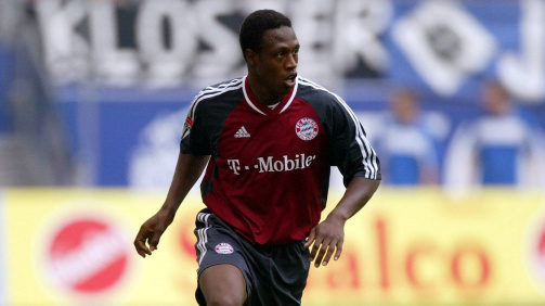 © imago - Fußballspieler Pablo Thiam in Aktion auf einem Rasenplatz. Er trägt ein Trikot des FC Bayern München mit den Farben Dunkelrot und Dunkelgrau sowie schwarze Stutzen mit roten Streifen (Saison 2001-02). Auf dem Trikot ist das Logo von T-Mobile als Sponsor zu sehen. Der Spieler ist in Bewegung und führt einen weiß-blauen Fußball mit dem rechten Fuß. Im Hintergrund sind unscharf weitere Personen sowie Werbebanner am Spielfeldrand zu erkennen. Die Szene spielt sich in einem Stadion ab.