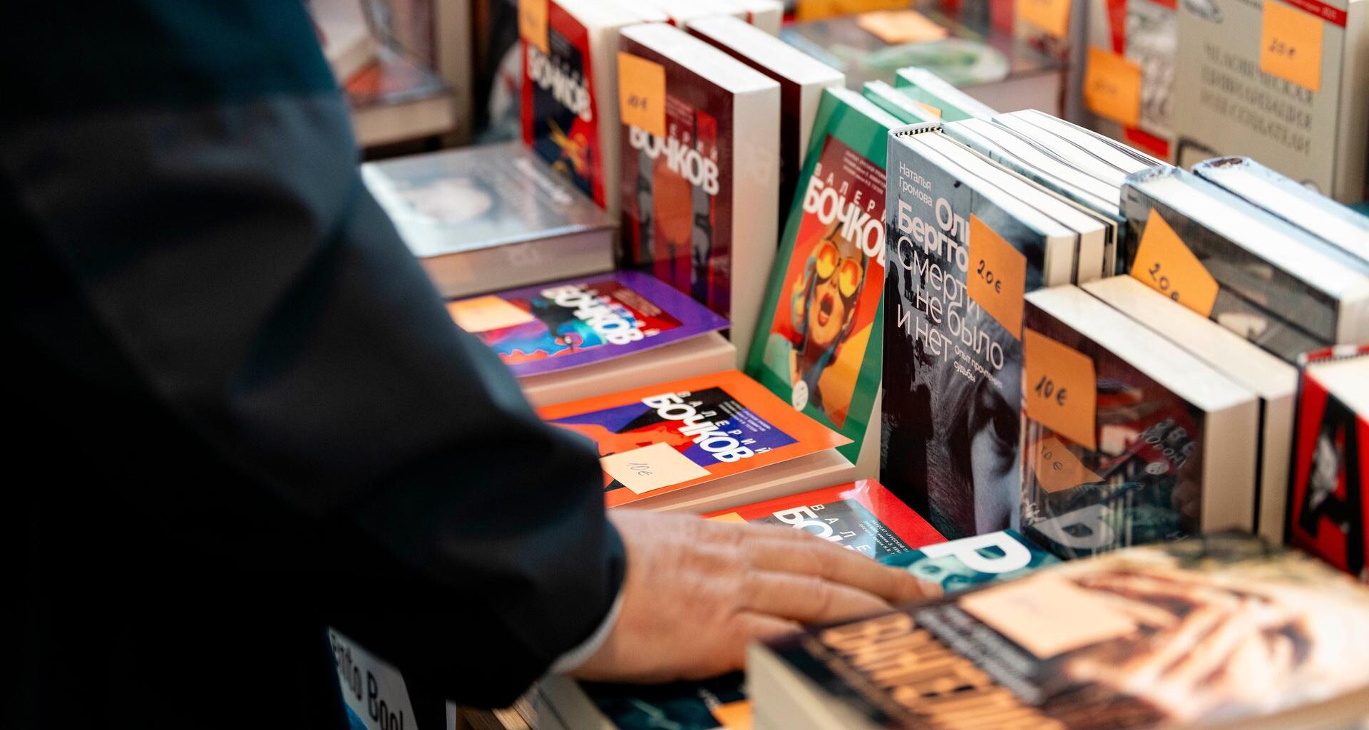 Auf der russischsprachigen Buchmesse in Berlin werden verschiedene russischsprachige Bücher zum Verkauf angeboten. Abgebildet ist ein Tisch, auf dem verschiedene Bücher aufgestellt und gestapelt liegen. Die Hand eines Mannes berührt eines der Bücher.